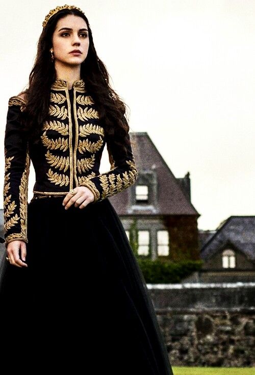 a young woman, possibly a princess or queen, stands in a black gown with gold embroidery. she is wearing a tiara and has long, dark hair. the dress is long and elegant, and the scene is set outdoors in front of a large mansion. the overall impression is one of historical royalty or fantasy.