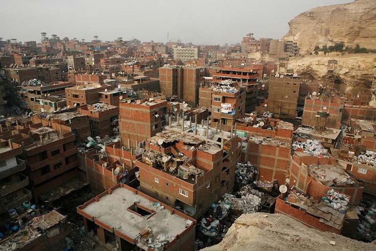 an aerial view of a densely populated slum area, possibly in cairo, egypt.  the image shows numerous closely packed, small, brick buildings in various states of disrepair.  rubbish and debris are visible throughout the settlement, piled high in some areas.  a large cliff face is visible in the background. the scene depicts extreme poverty and overcrowding. satellite dishes are visible on many of the buildings, suggesting a degree of access to technology despite the impoverished conditions.