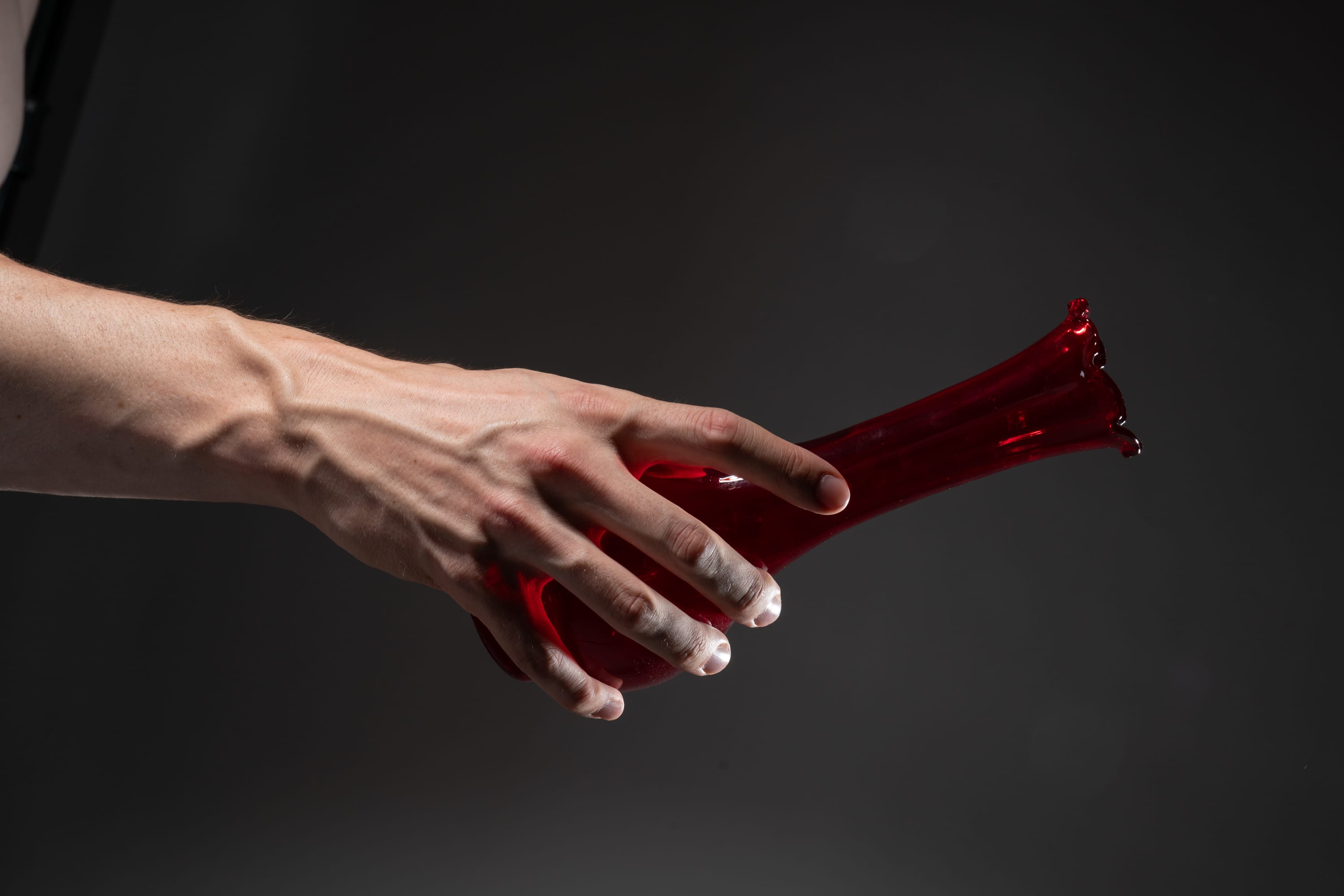 a male hand is holding a red glass vase against a dark gray background. the hand is positioned in a way that highlights the veins and muscles. the image has a studio lighting setup.