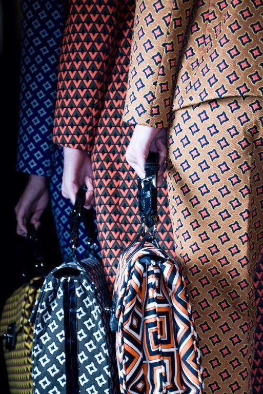 loseup shot showcasing a group of models at a fashion show, each wearing distinct patterned suits with geometric designs. the suits feature a mix of vibrant colors including orange, blue, black, and white, with intricate geometric patterns that vary slightly between each model.  each model is carrying a matching patterned handbag, with the bags also featuring a geometric design. the focus is on the detail of the fabrics and patterns.  the overall impression is one of stylish, sophisticated autumn/winter fashion.