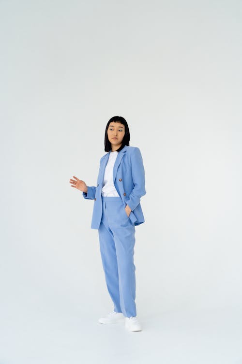 a fullbody shot of a young asian woman in a light blue pantsuit standing against a white background. she's wearing a white shirt underneath and white sneakers. her posture is confident, and she's making a subtle hand gesture, possibly indicating something. the overall style is minimalist and elegant.