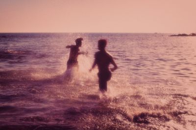 a vintagestyle photograph depicting two young men silhouetted against a sunsetlit ocean as they playfully splash in the water. the scene is peaceful and evokes feelings of summer, friendship and carefree fun.  the image has a slightly hazy, nostalgic quality consistent with older film photography.