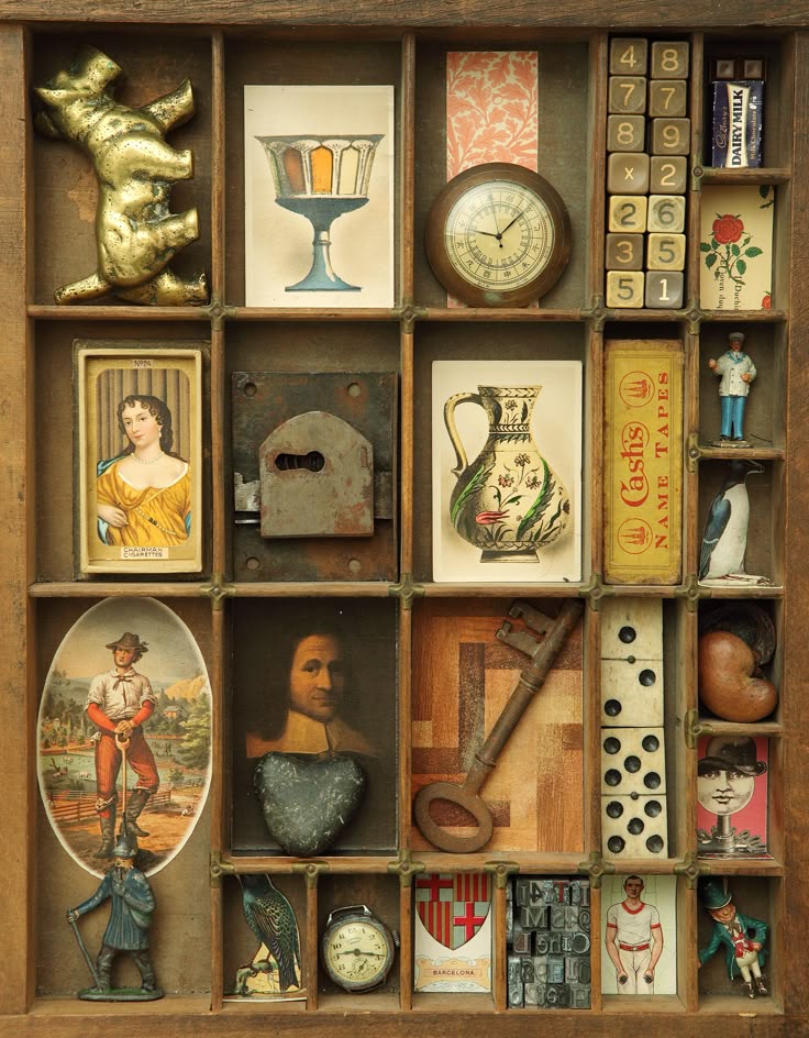 a wooden box with compartments filled with a collection of vintage objects, prints, and ephemera. the arrangement includes framed images, clocks, keys, figurines, dominoes, cards, locks, and other collectible items, showcasing a nostalgic assemblage.