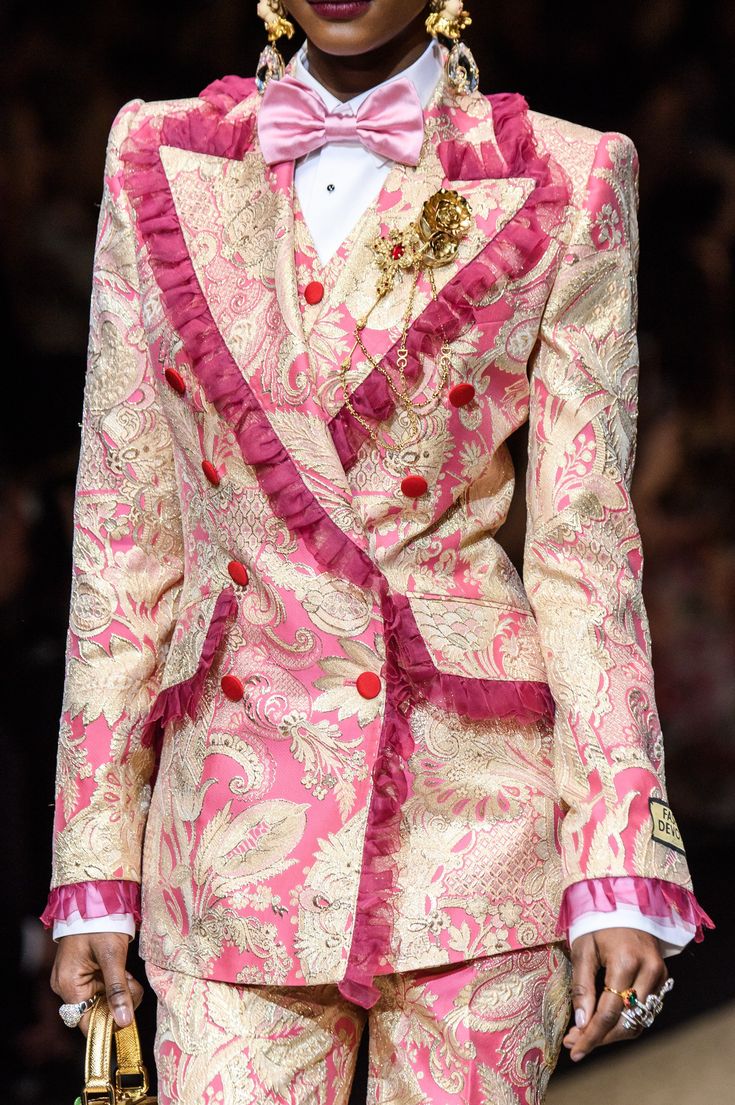 closeup shot of a model showcasing a dolce & gabbana runway outfit. the model is wearing a pink and gold brocade suit featuring a paisley and floral pattern.  the suit is a doublebreasted jacket and trousers, trimmed with pink ruffles.  the model also wears a pink bow tie, gold jewelry including statement earrings and a brooch, and several rings. the image focuses on the intricate detail and luxurious texture of the clothing.