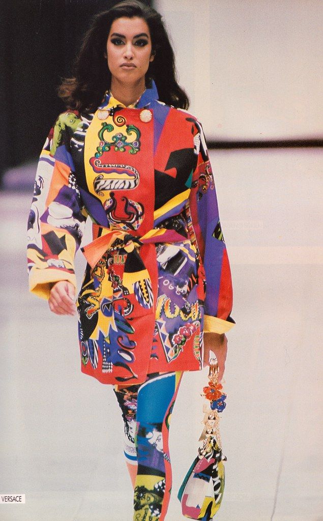 a fashion model walks the runway wearing a colorful versace ensemble consisting of a jacket and matching pants. the jacket is belted at the waist and features a vibrant print design with various shapes, colors, and patterns. the pants also have a matching print. the model is carrying a handbag adorned with colorful embellishments. the style appears to be from the 1990s, giving it a vintage aesthetic.