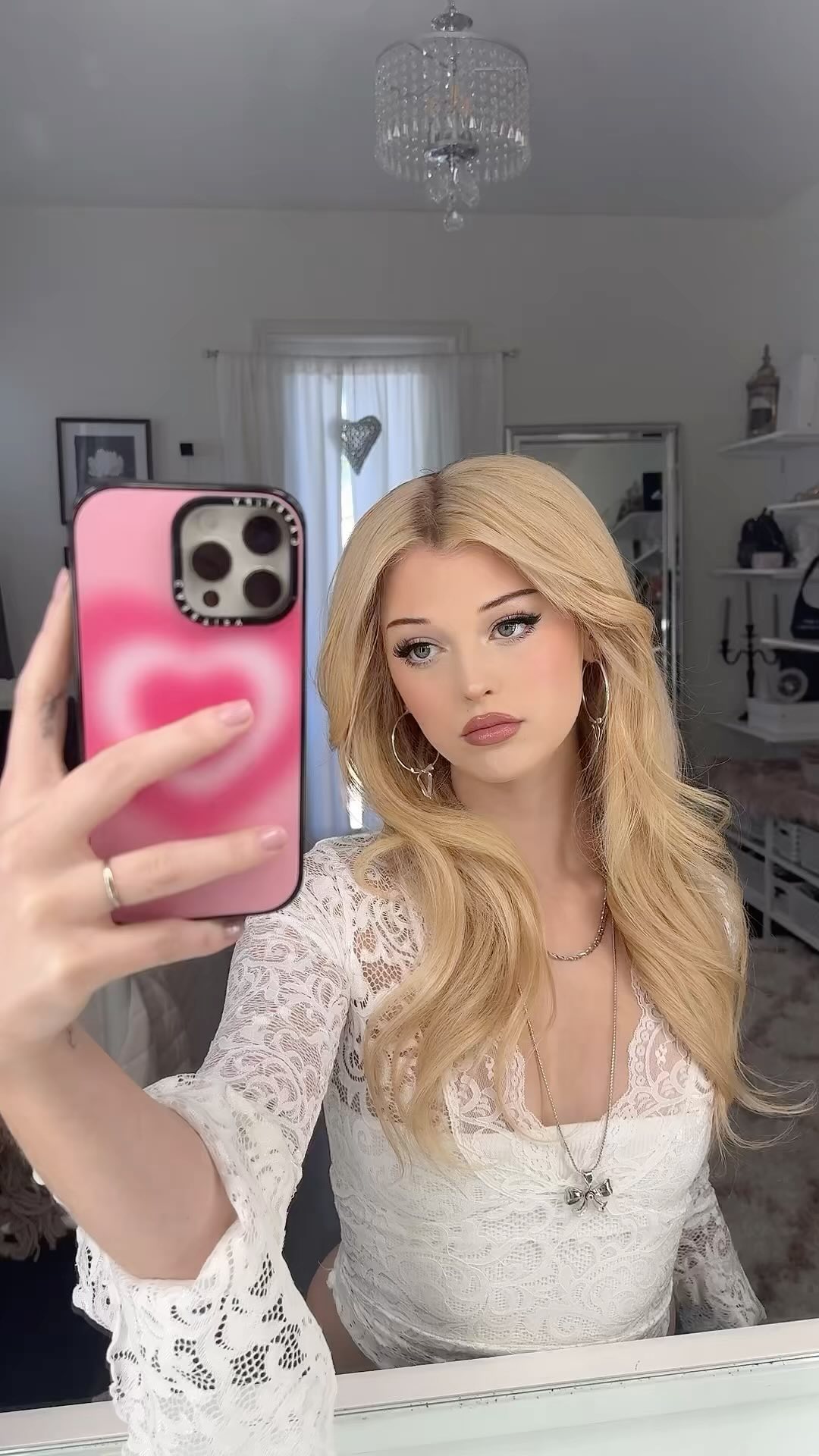 a young woman with long blonde hair takes a selfie in a mirror. she is wearing a white lace top with long sleeves, hoop earrings, and a necklace. she is holding a pink phone with a heart design. behind her, a chandelier hangs from the ceiling, and a mirror reflects the room. the room appears to be a bedroom with a decorated shelf and other furniture in the background.