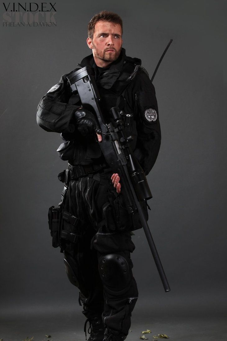 a soldier in full military gear stands with a rifle in hand, looking to the right of the frame. he is dressed in black camouflage and is holding a sniper rifle with a scope. the background is a plain gray.