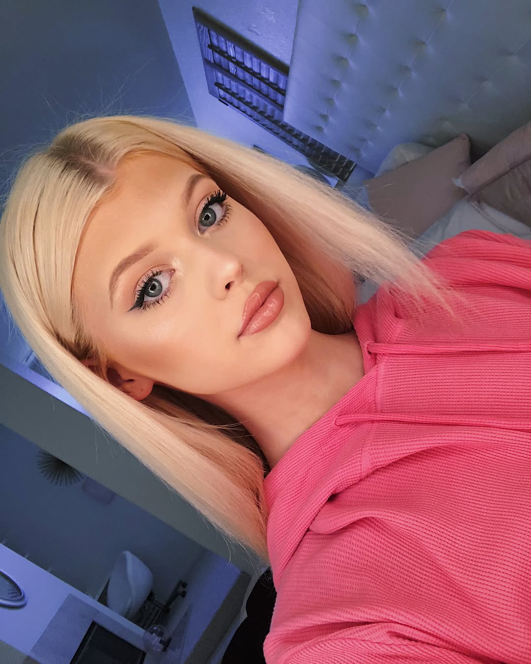 a closeup selfie of a woman with blonde hair wearing a pink hoodie. she has makeup on, including eyeliner and lipstick. she is indoors, likely in a bedroom, as there is a bed, pillows, and a headboard in the background. there are also blinds visible in the background. she is posing and looking at the camera.