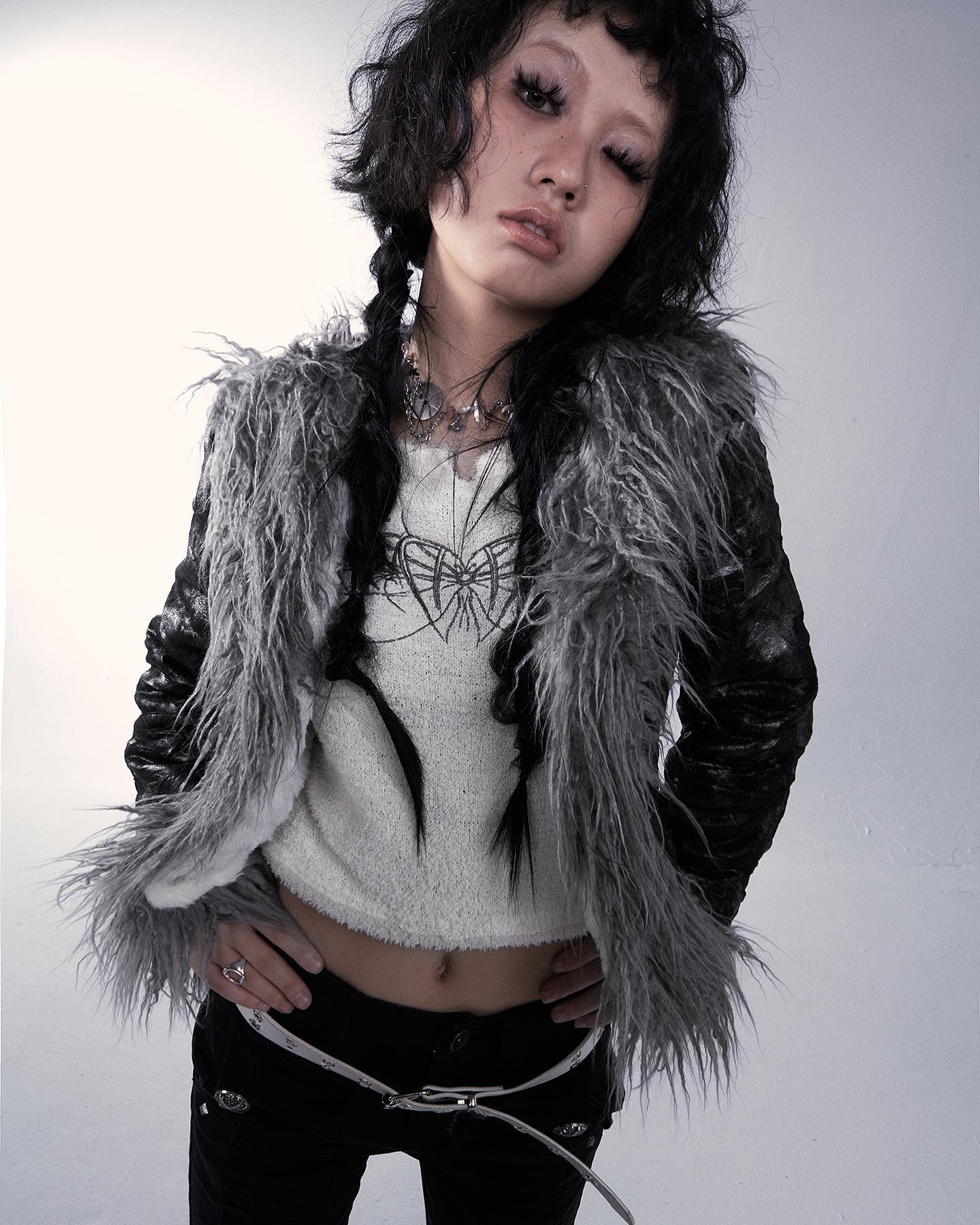 the image is a fashion portrait of a young woman. she's posing in a studio against a plain white backdrop. her style is edgy and alternative, featuring a gray faux fur jacket over a white cropped sweater and black pants. she is wearing a silver belt. her dark hair is styled in a partially braided look with bangs. her makeup is dark and emphasizes her eyes. she is looking directly at the camera with a slightly serious, cool expression. the overall aesthetic is gothic or grungeinspired.