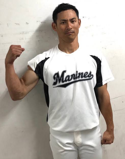a japanese baseball player, wearing a white jersey with black lettering that reads "marines," stands with his right arm flexed and a fist, smiling happily. he's wearing white baseball pants and is standing against a white wall.