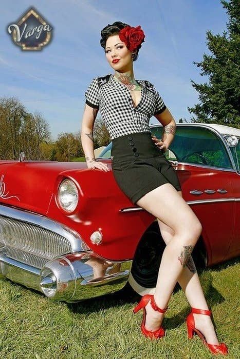 a beautiful pinup girl dressed in a vintage outfit is posing next to a red classic car. she is wearing a black and white checkered shirt with a black skirt and high heels. she has red lipstick and her hair is styled in a victory roll with a red flower.  she has a tattoo on her arm.