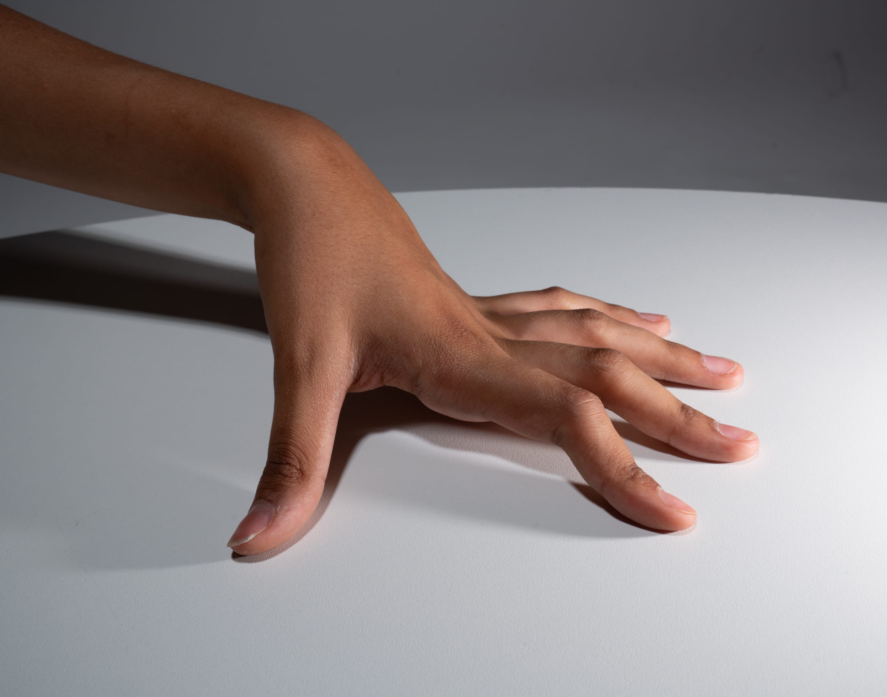 a closeup studio shot of a person's hand with brown skin reaching across a white table. the lighting is bright and casts a distinct shadow of the hand on the surface. the focus is on the fingers and their position as they gently touch the table.