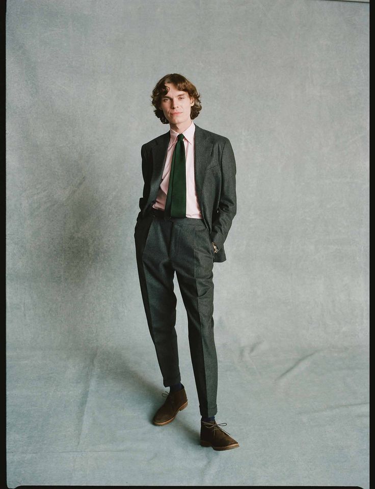 a full shot of a young man, model, standing against a gray textured backdrop. he's elegantly dressed in a dark gray suit paired with a light pink shirt and a dark green tie. his shoes are brown suede desert boots. the overall style is a smart casual blend of formality and relaxed elegance. the image has a clean, sophisticated feel thanks to the muted color palette and careful styling. the model is wellgroomed and projects a confident and stylish image.