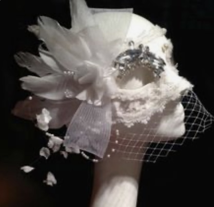 a white masquerade mask with intricate lace detailing is displayed on a stark white mannequin head against a black backdrop. the mask is adorned with delicate white feathers, shimmering beads, and sheer white tulle, creating a dramatic and elegant effect. a small cluster of beads sits on the upper right side of the mask, adding a touch of sparkle. the soft, diffused lighting emphasizes the delicate textures and intricate details of the mask. the overall impression is one of refined beauty and mystery.