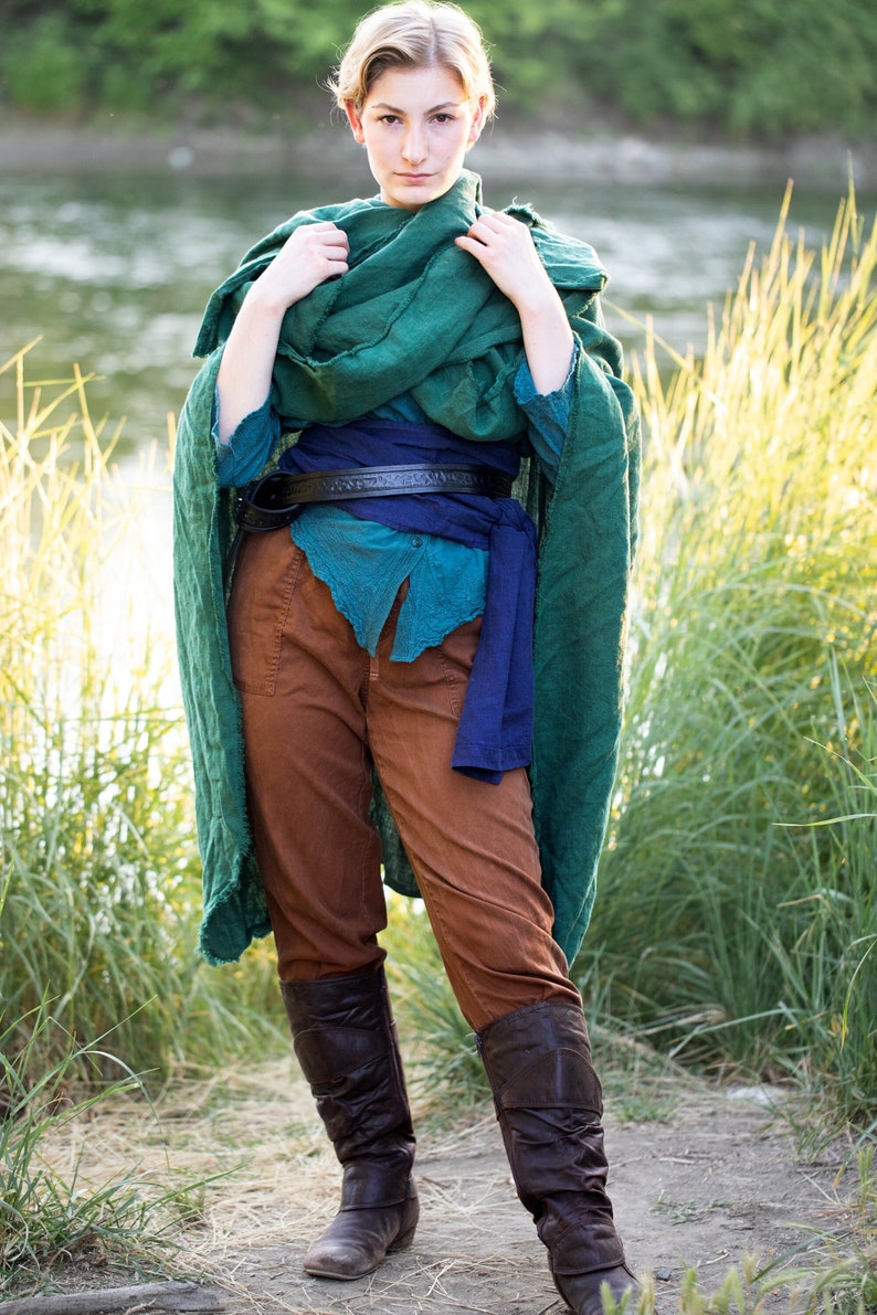 a woman is standing outdoors wearing brown pants, boots, a teal shirt, a dark blue sash, a black belt, and a green shawl. she is standing in front of water and tall grass.