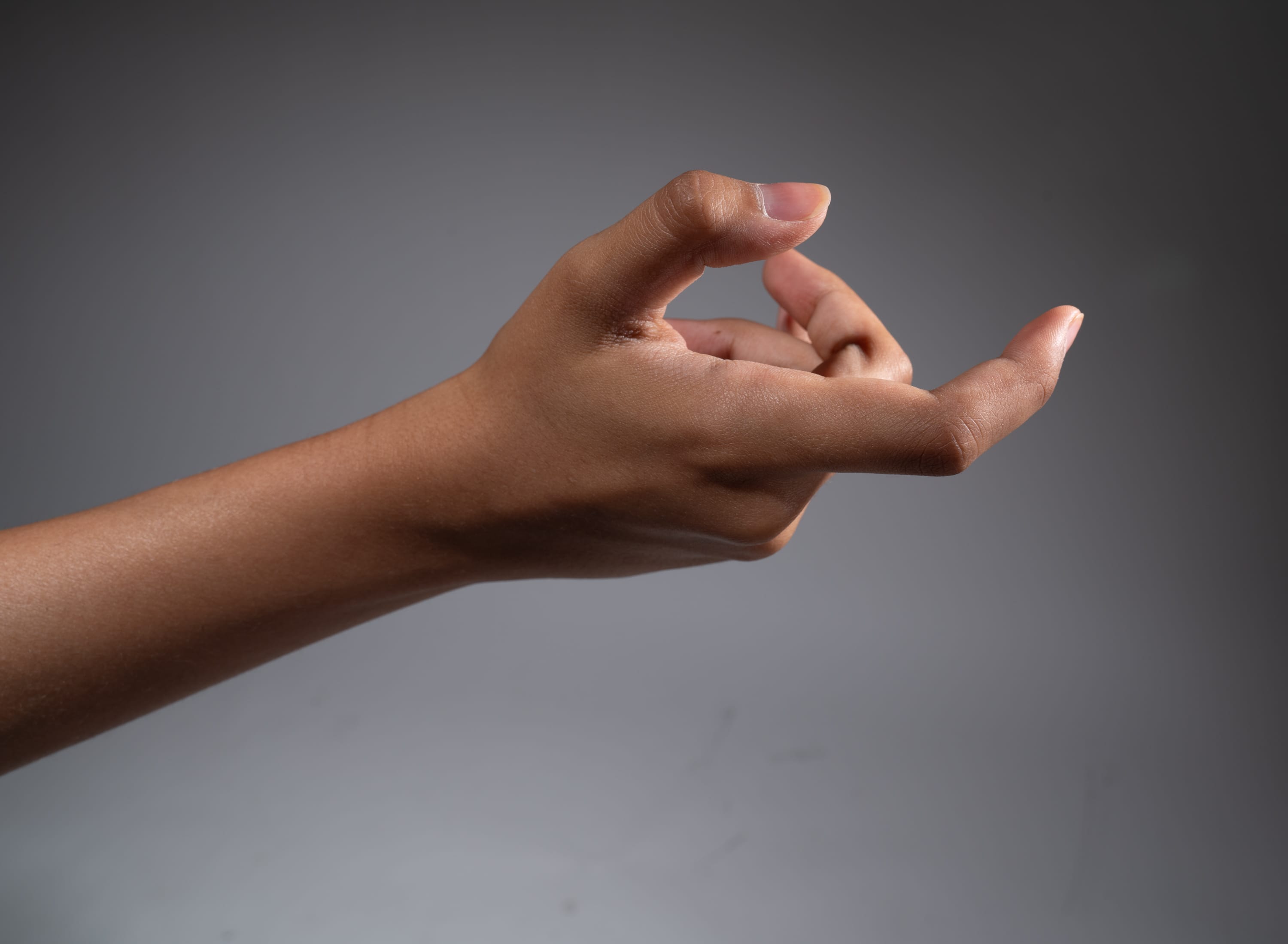 a hand is reaching out with the index finger and thumb pinching together as if picking something up. the hand is positioned against a plain gray background, illuminated by studio lighting.