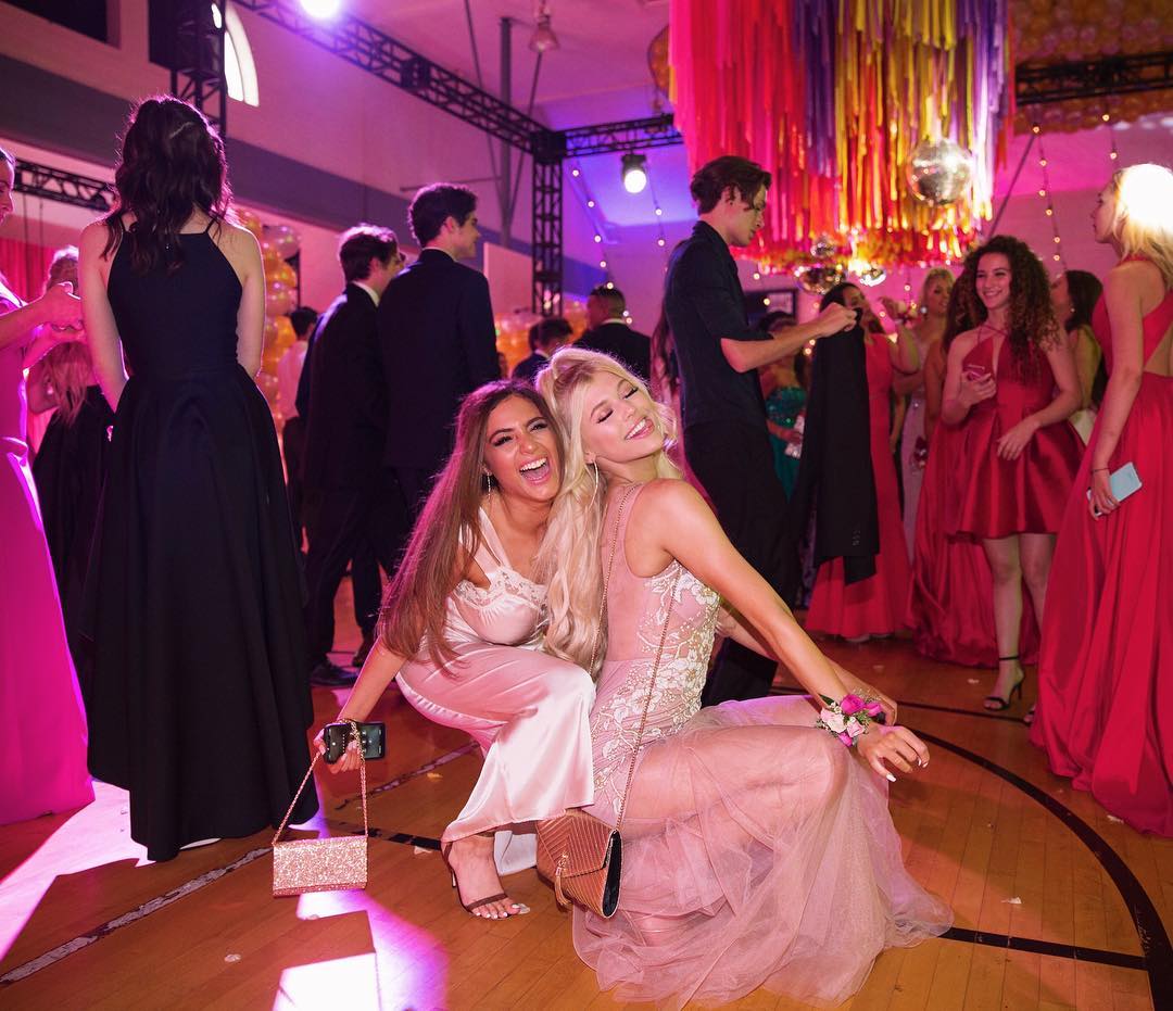this image shows a group of young women and men dressed in formal attire at a prom or similar formal dance. two girls are crouching in the foreground, smiling and seemingly enjoying themselves. the background is filled with other attendees, decorations, and a disco ball hanging from the ceiling. the scene is lively and joyful, capturing the essence of a memorable social event for high school students.
