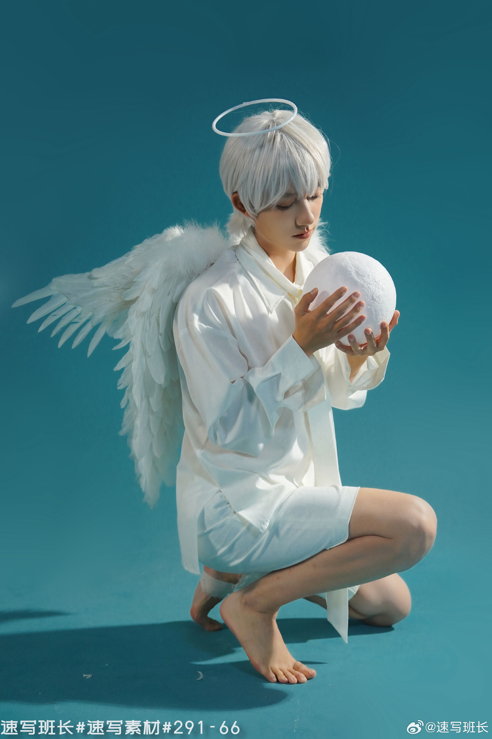a young man in white is posing against a light blue background. he has white hair and is wearing white clothing and white angel wings. he also has a halo above his head. he is barefoot and crouching down while holding a white orb. the photo is captioned with chinese characters.