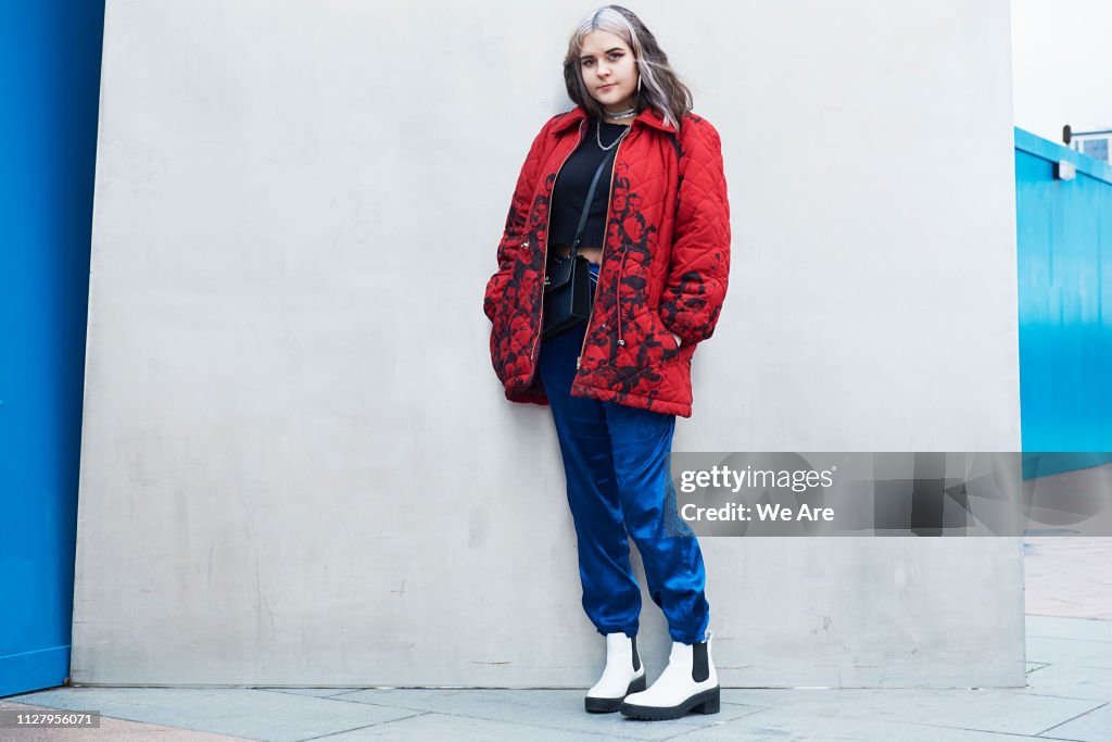 a woman in street style fashion stands against a wall. she's wearing a red puffer jacket with black floral print, blue pants, and white boots. she also has a black top and a crossbody bag. her hair is silver, and she wears a silver necklace.