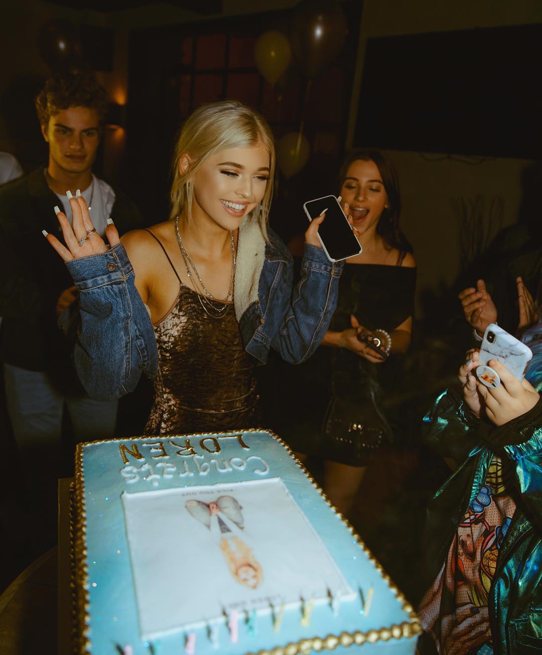 a smiling woman stands in front of a birthday cake that says "congrats loren," holding her hands up in celebration. she is wearing a shimmery dress and a denim jacket. surrounding her are other people, including a man in the background and two women, one of whom is holding a phone. there are balloons in the background and a birthday cake in the foreground. people are taking pictures with their phones.