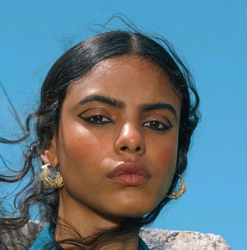 a closeup portrait of a young woman with long, dark, wavy hair blowing in the wind. she has tan skin and is wearing winged eyeliner and gold leafshaped hoop earrings.  her expression is serious and intense. the background is a clear blue sky and a partially visible stone surface. the overall mood is one of beauty and fashion.