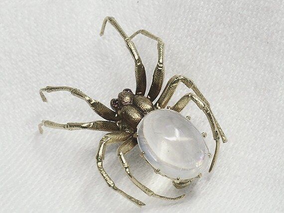 closeup of a silver and gold spider brooch with a clear crystal gemstone in the center. the spider is detailed with small red gems for eyes and is set against a white background. the brooch appears to be crafted from metal with intricate details.  the clarity of the gemstone and the fine craftsmanship of the spider create a luxurious and slightly spooky effect.