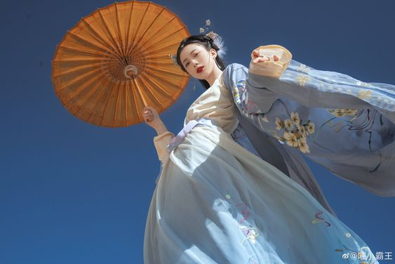 a woman in a flowing, light blue hanfu stands against a vivid blue sky. she holds a traditional bamboo umbrella, its golden color complementing her attire. the intricate embroidery on her clothing, featuring floral motifs, adds a touch of elegance. the lowangle perspective emphasizes the grace and beauty of both the woman and her traditional garb.