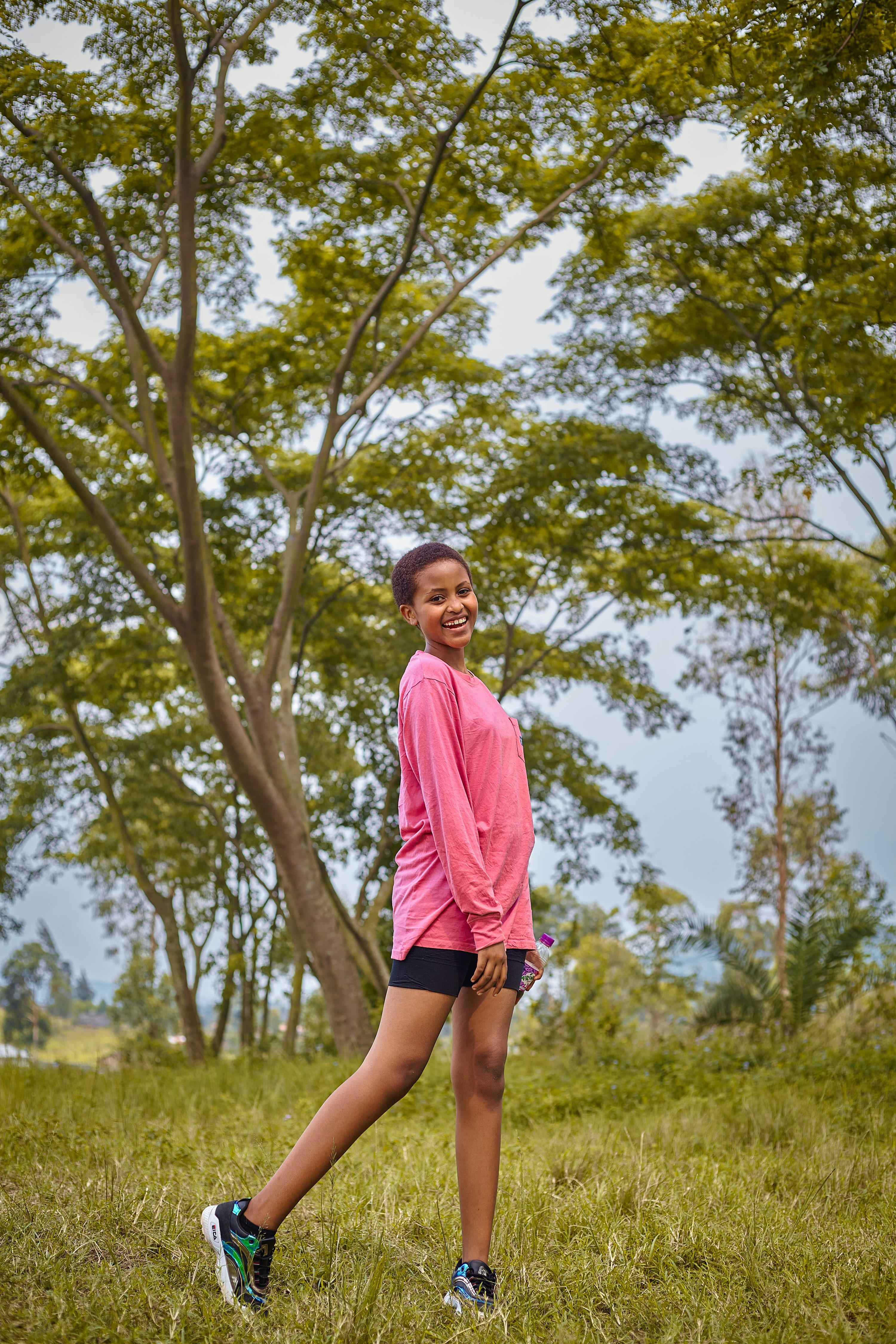 a young african woman with short hair is walking through a park, smiling happily. she is wearing a pink longsleeved shirt, black athletic shorts, and colorful sneakers. the setting is outdoors on a sunny day, and the background is filled with lush green trees and grass.  she appears to be enjoying a healthy and carefree outdoor activity.