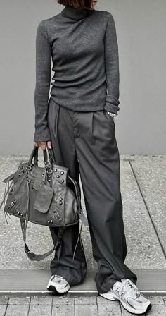 a woman is standing on a grey sidewalk, with a grey sweater and black pants. she is carrying a grey handbag over her shoulder and wearing white sneakers. she has a fashionable look and is posing for the camera.