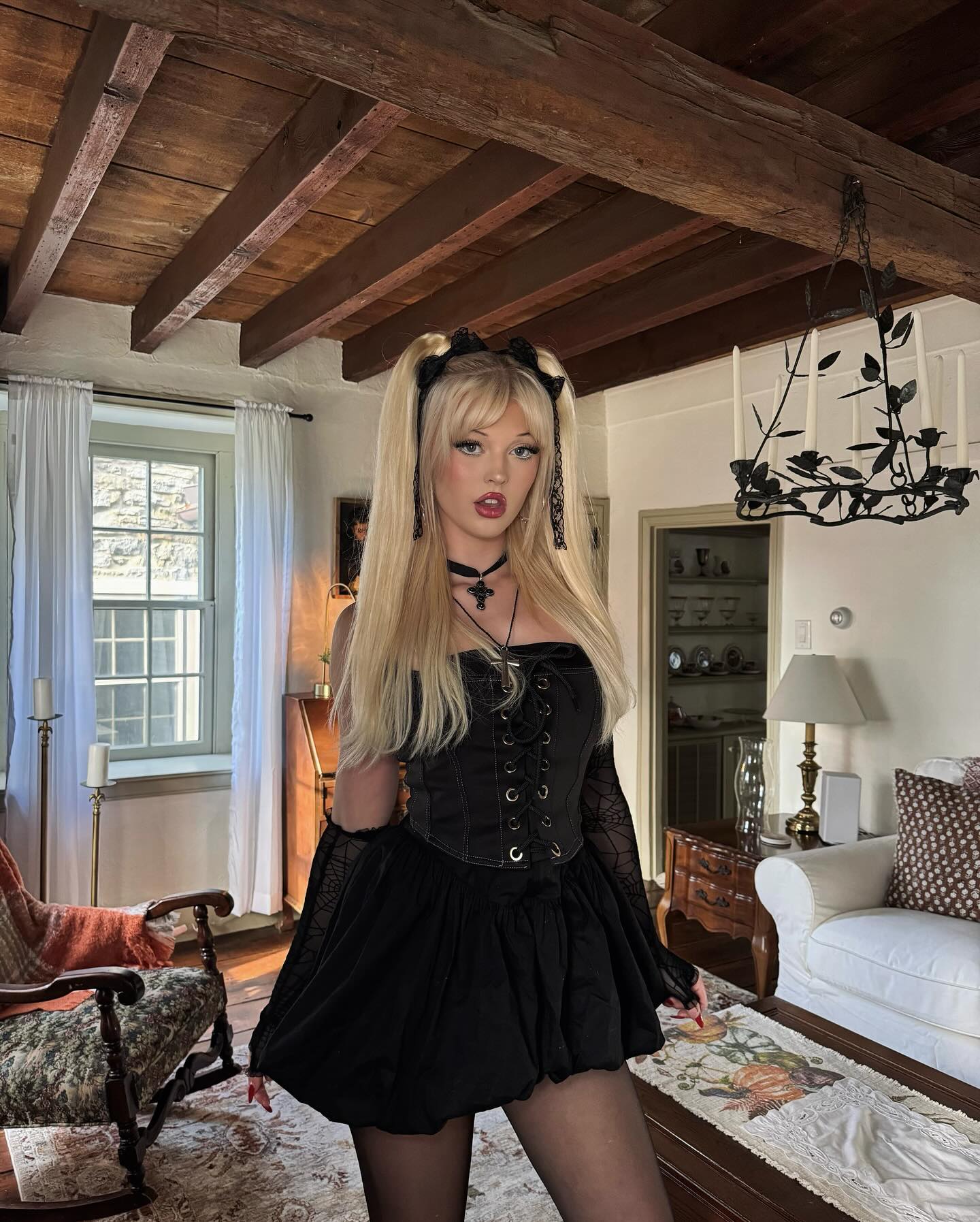 a young woman with blonde hair styled in pigtails, adorned with black ribbons, stands in an interior setting. she is wearing a strapless black dress with a corsetstyle bodice and a short, flared skirt. she also sports a black choker necklace with a cross pendant and black tights. the room features a mix of antique and modern elements, including wooden beams, a vintage armchair, a white sofa, a decorative chandelier, and a window providing natural light. the overall aesthetic suggests a gothic or alternative style.