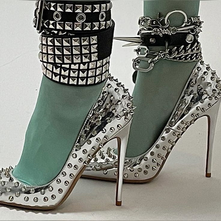 a pair of silver high heels with a pointed toe and a stiletto heel. the shoes are covered in spikes, giving them a punk rock edge. they are also adorned with a silver chain and buckle around the ankle. the shoes are elegant and glamorous, but also edgy and trendy. they would be perfect for a night out on the town or a special occasion.