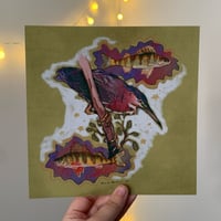 loseup view of a hand holding a vibrant artwork print. the print features a green heron perched on a branch, with two perchlike fish depicted in stylized, almost dreamlike clouds surrounding the bird. the color palette is rich, with greens, purples, and golds dominating the piece. the style is illustrative, possibly a mixedmedia painting, with a slightly surreal or whimsical feel to it. the background is a muted olive green. the image is welllit, and the details in the artwork are clearly visible.