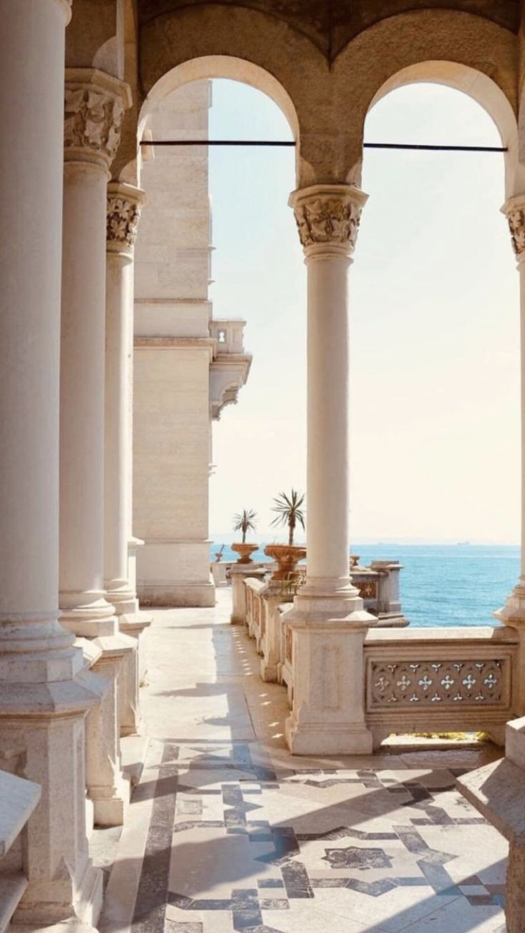 the image depicts a sunlit, elegant colonnaded terrace or balcony overlooking a calm sea. the architecture is classical, featuring tall, slender marble or stone columns supporting graceful arches.  intricate details adorn the columns and balustrade. the floor is tiled with a geometric pattern. two small palm trees are visible in planters on the terrace, adding a touch of nature to the scene.  the overall impression is one of serenity and luxury. the color palette is predominantly warm, cream, and beige tones, enhanced by the bright sunlight.