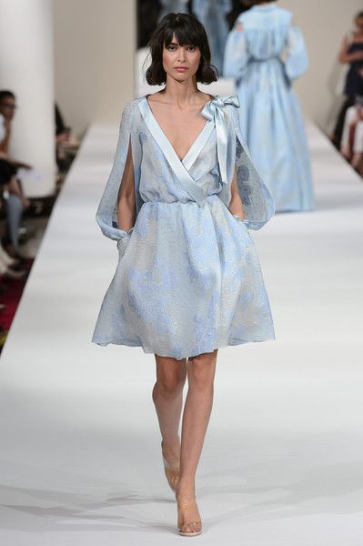 a fashion model walks the runway wearing a light blue, aline dress with cape sleeves and a delicate bow detail at the neckline. the dress has a subtle textured fabric and appears to be embroidered. the model has shoulderlength dark brown hair and is wearing clear high heels. the runway is white and other models can be seen in the background, all wearing similar pastelcolored garments. the overall atmosphere is elegant and sophisticated, suggesting a highend fashion show.
