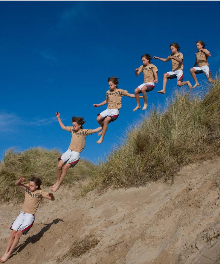 a group of boys, with tan skin, dressed in tan shorts and shirts, jump and play in the sand dunes. they are smiling, enjoying their day in the sun and having fun with their friends.