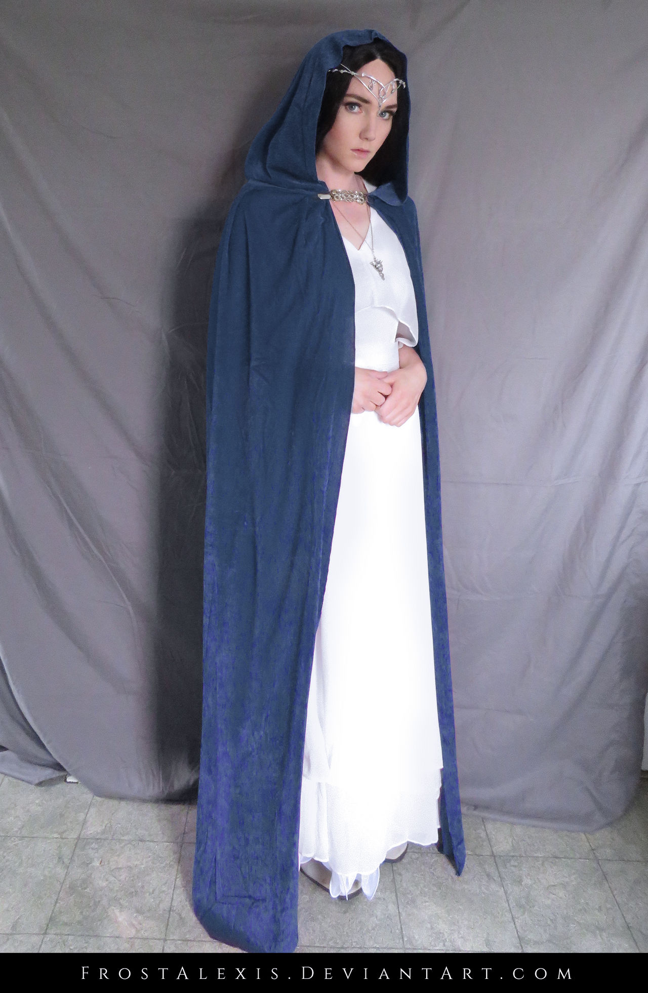 a fullbody shot of a woman cosplaying as an elven character. she's wearing a long, dark blue hooded cloak over a flowing white dress.  she also wears an elvenstyle tiara and necklace. the background is a simple gray backdrop.  the image is welllit and professionally taken, suggesting it may be from a cosplay portfolio or photoshoot. the watermark "frostalexis.deviantart.com" is visible at the bottom.