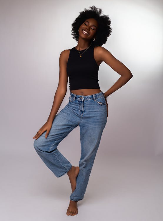 a fullbody studio shot of a smiling, barefoot young black woman with a large afro. she is wearing a black crop top, light blue denim jeans, and a necklace. her right leg is planted, and her left leg is lifted with her foot resting on her right knee. her left hand is resting on her right thigh, and her right hand is on her right hip. she is facing forward but slightly turned to the left, looking up with her eyes closed.