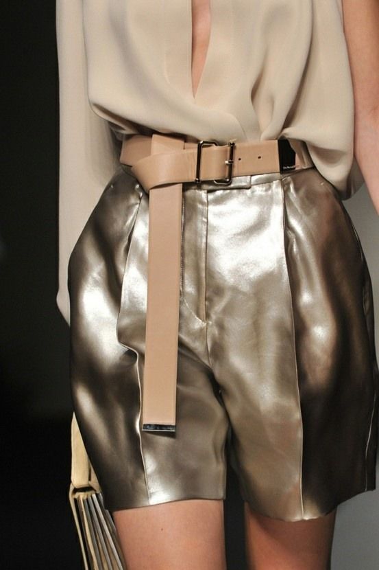 a model wearing a silver metallic shorts and a beige top with a silver belt. the model is posing on a runway with her legs and body in a flattering pose. she is wearing her hair down and her makeup is minimal. she is giving off an air of confidence and style.