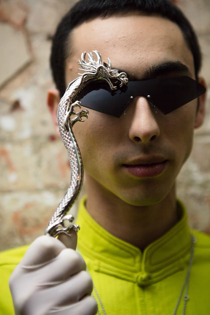 a man with dark hair is wearing angular black sunglasses and a bright yellow shirt. he is holding a silver dragon ornament in his gloved hand. the head of the dragon is positioned near his face. a silver chain necklace is visible on his chest. the background appears to be a textured wall.