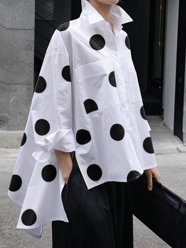 a woman wearing a long white polka dot shirt with black dots and black pants poses for a photo. she's holding a black clutch purse and has her hair pulled back.