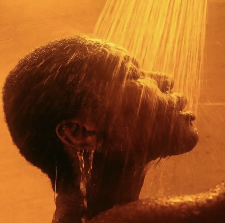 the image is a closeup, artistic shot of a darkskinned person's head and upper body under a shower. the water streams are intensely lit with a warm, amber or sepiatoned light, giving the overall scene a very cinematic and sensual mood. the focus is on the individual's face and the contrast between their dark skin and the flowing golden water. the lighting and color palette evoke feelings of relaxation, warmth, and peace. the image has a very stylized and aesthetic quality, possibly taken from a film or artistic project.