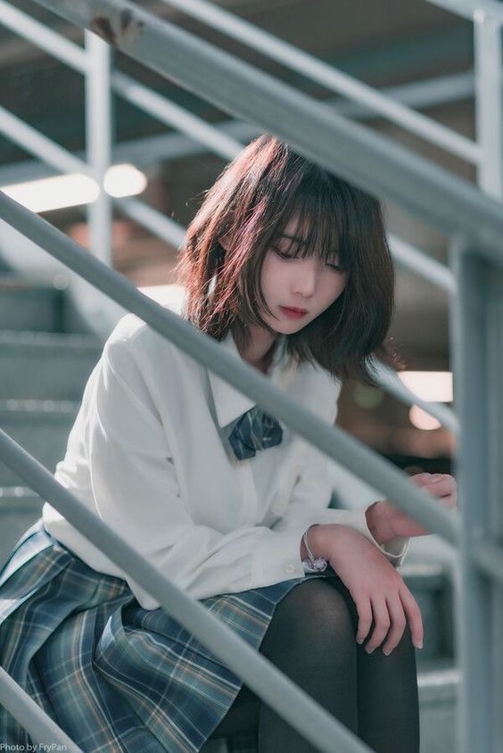 a young girl in a school uniform sits on the stairs with a sad expression. she is wearing a white shirt, plaid skirt, and black tights. her hair is covering her face and she looks lonely.  the image has a melancholy tone.