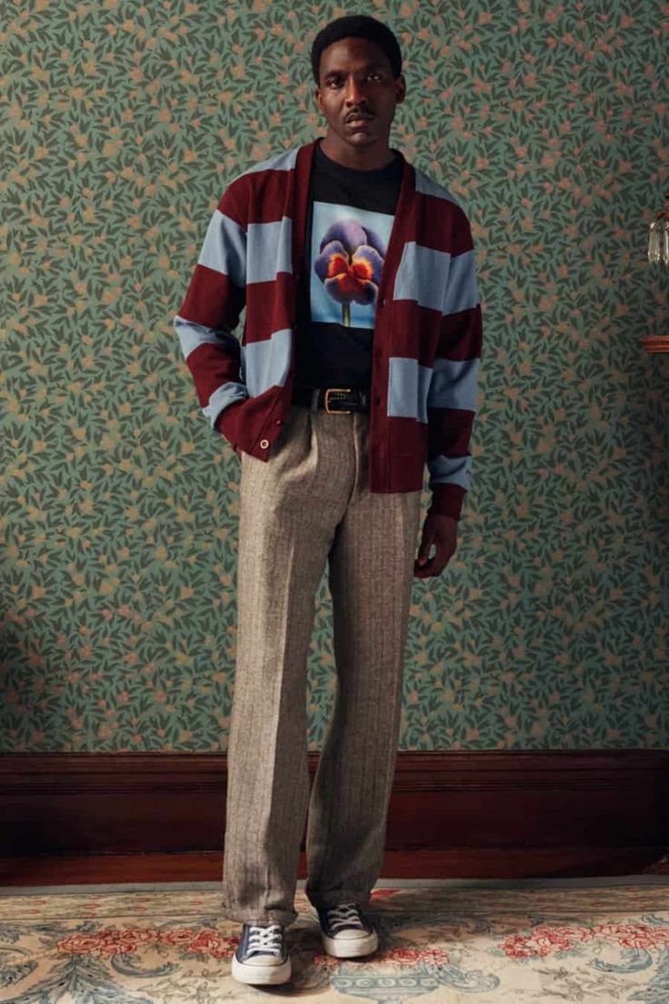 a fullbody shot of a male fashion model standing against a vintage wallpaper backdrop. he is wearing a burgundy and light blue striped cardigan over a black tshirt with a floral print, brown herringbone pants, and black converse sneakers. he has a belt around his waist and a serious expression on his face. the image is welllit and professionally styled.
