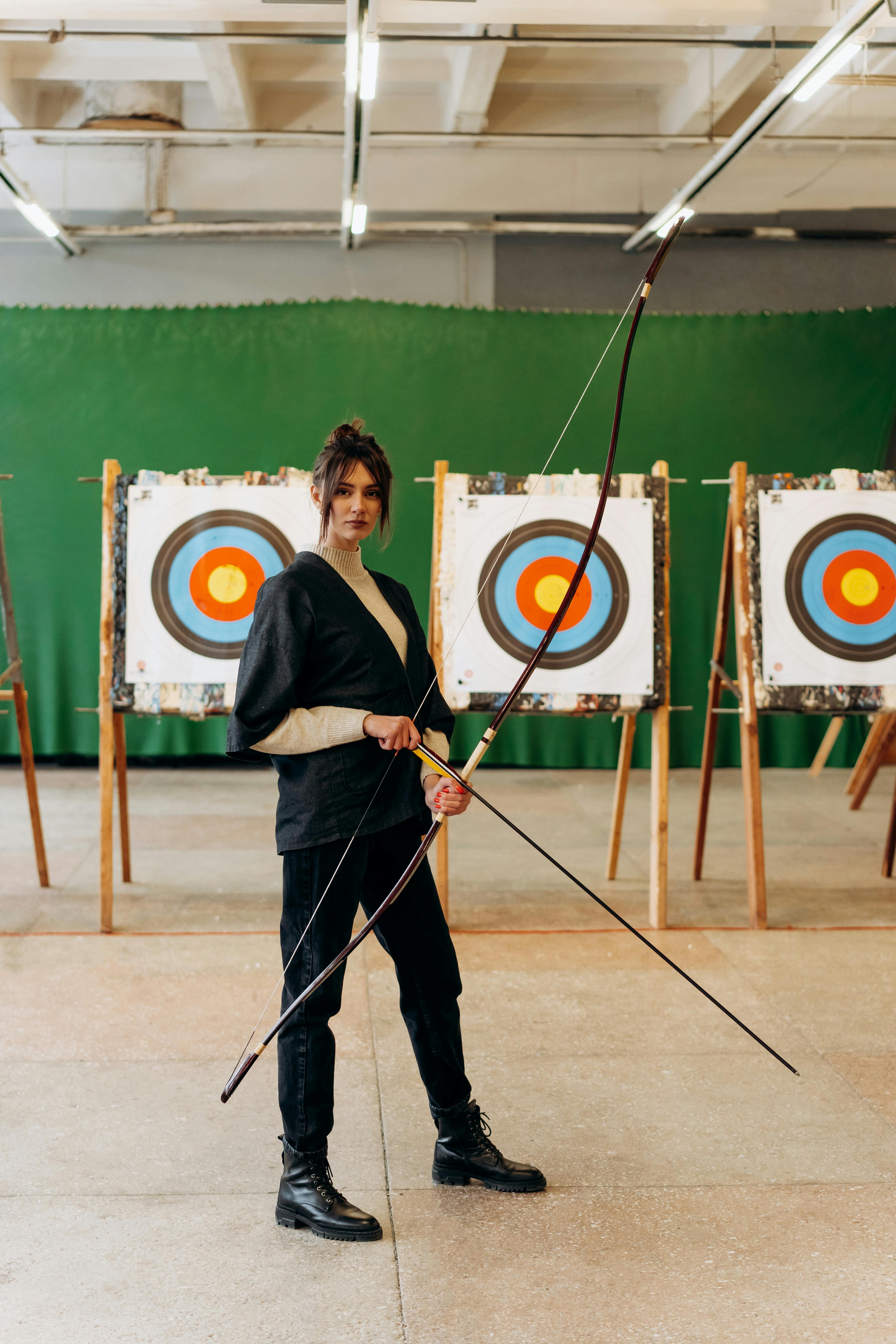 a young woman stands confidently in an indoor archery range, holding a bow and arrow. she is dressed in black clothing and boots and is focused on the target in front of her.  three target stands are visible, all displaying archery targets. the background is a green wall. the overall mood is one of concentration and athleticism.