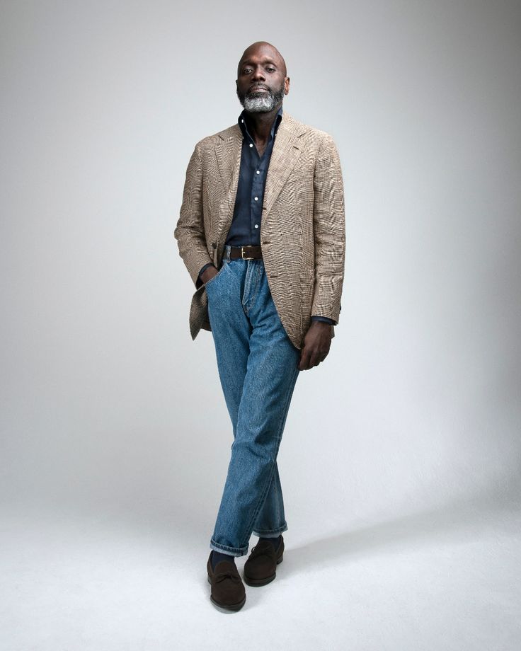 a fullbody studio shot of an older african american man with gray hair and a beard. he's wearing a beige tweed sport jacket, a dark blue buttondown shirt, blue straightleg jeans, and brown loafers. his expression is serious, and he's standing confidently against a neutral gray background. the overall style is smart casual.