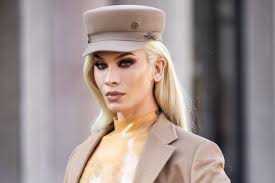 the image shows a closeup portrait of a blonde drag queen or transgender woman. she's wearing a stylish beige newsboy cap, a tan blazer, and a gold, possibly latex, top.  her makeup is impeccable, and her expression is serious and composed. the background is blurred, suggesting an outdoor setting. the overall impression is one of elegance and sophistication.