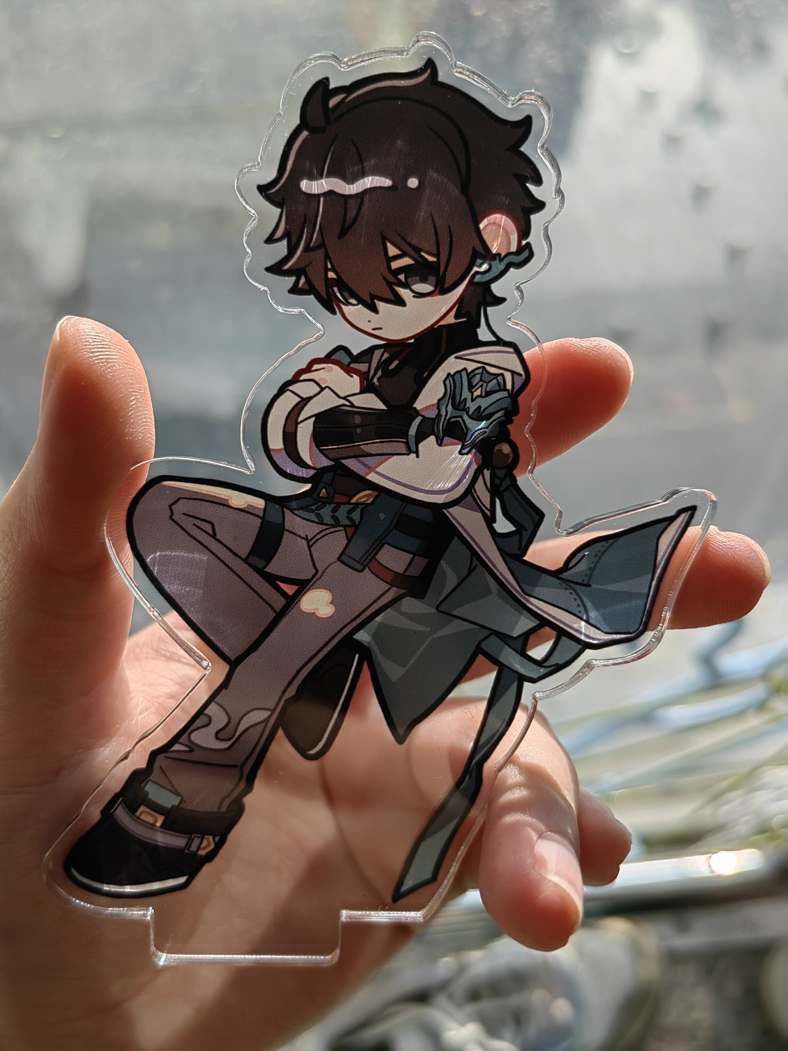 a hand is holding an acrylic stand of an anime character. the character is a young boy with dark hair and a serious expression. he is wearing a lightcolored coat and pants and is posing with his arms crossed. the stand is clear and has a white base. the character's design is based on a popular anime or manga.