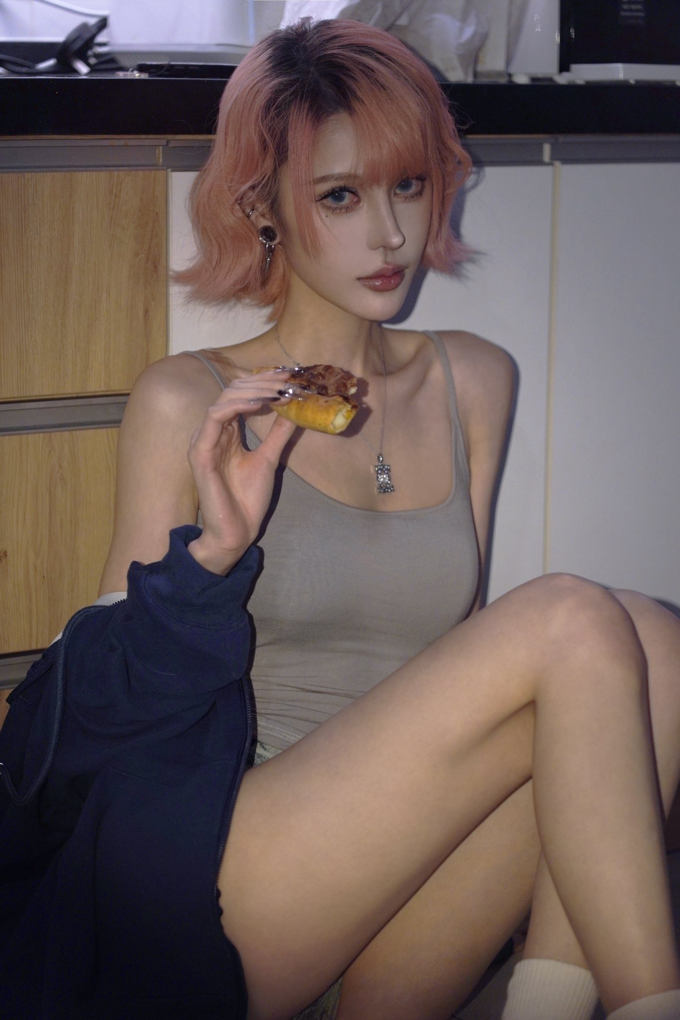 a young woman with short, pink hair is sitting in what appears to be a kitchen. she is wearing a grey tank top and a dark blue jacket. she is holding and eating a small pastry.  she has a necklace and earrings on. the image is a portrait shot, focusing on the woman. her expression is calm and relaxed.