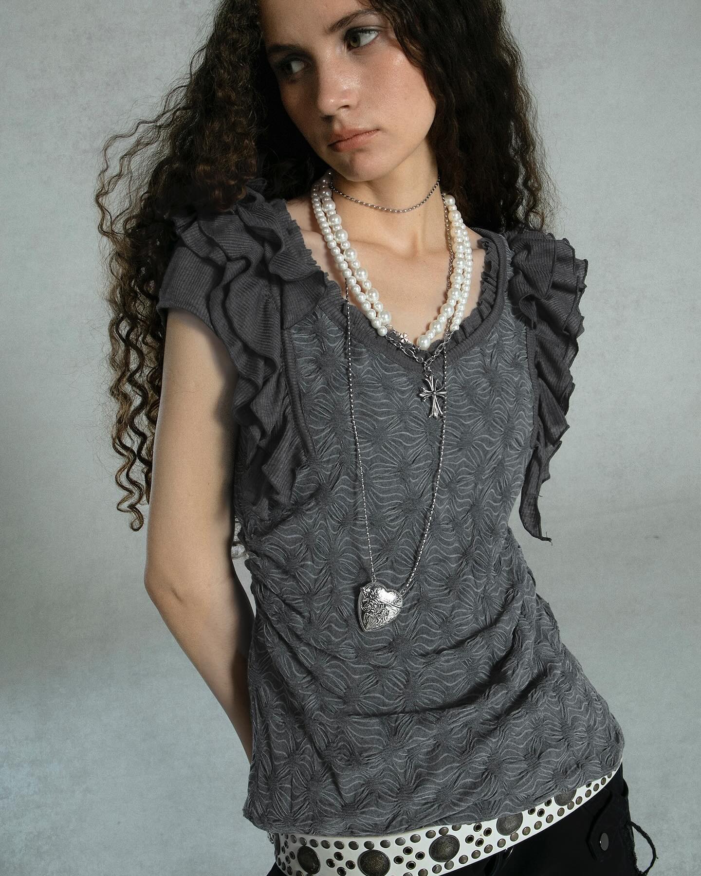 a young woman with long, curly brown hair is modeling a gray ruffled top. the top is textured and has a slightly gothic aesthetic. she is wearing a doublestrand pearl necklace and a silver necklace with a cross and a heart pendant. the woman is posing against a gray backdrop. the overall style is elegant yet alternative.
