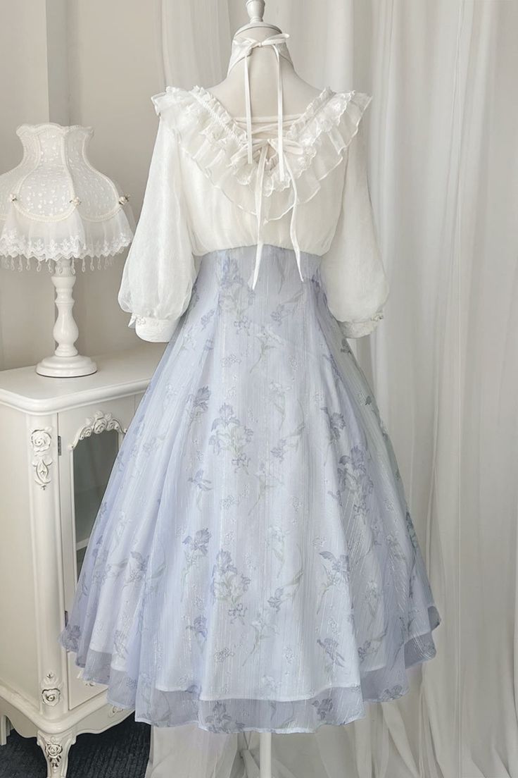 a white lace blouse and a flowing blue floral skirt are displayed on a mannequin. the blouse features delicate lace detailing on the neckline and sleeves, while the skirt boasts a full, romantic silhouette with intricate floral patterns. the soft, natural lighting highlights the textures and details of the garments, creating a dreamy and elegant aesthetic.